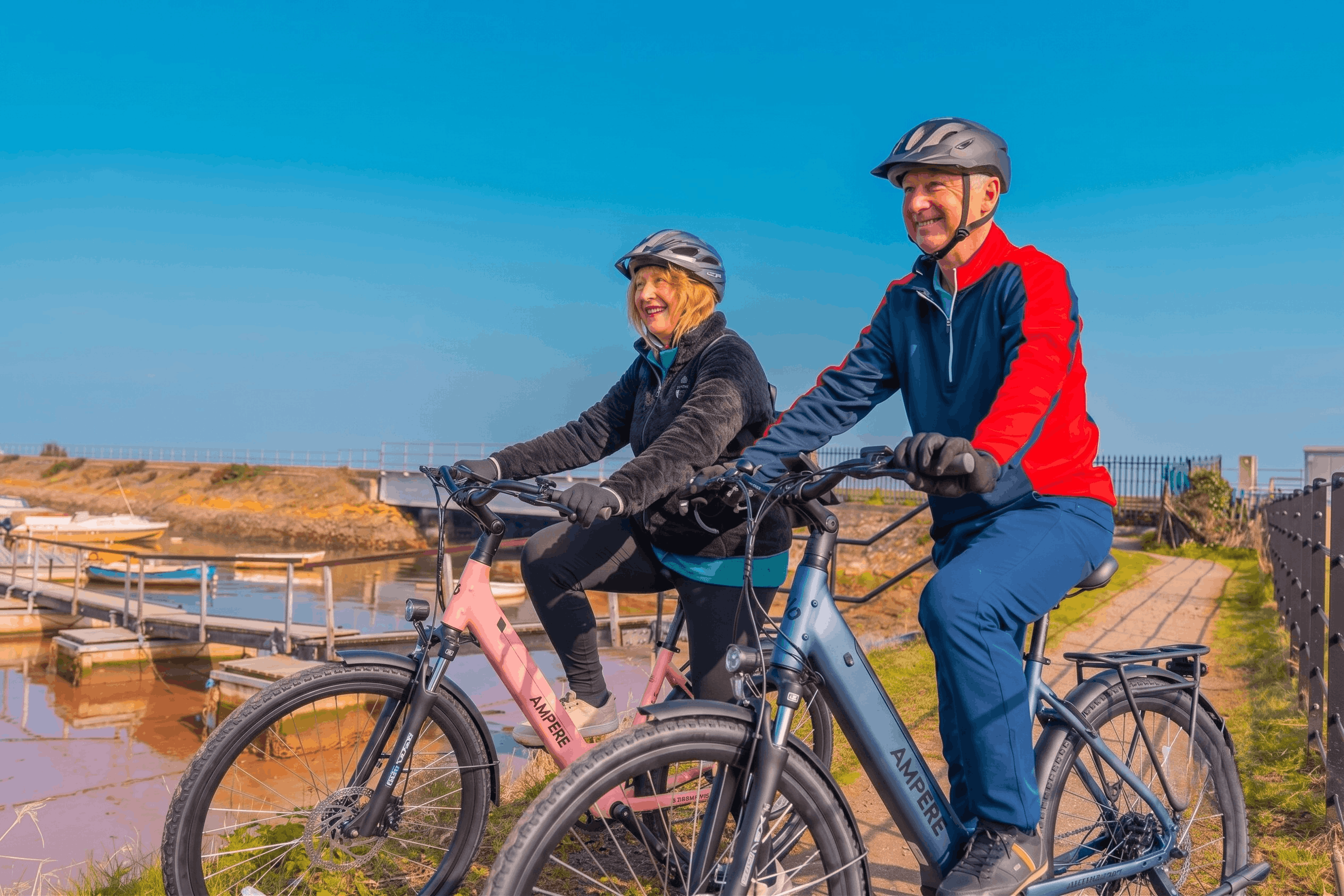 Pink Ampere Deluxe 700 and Stone blue Ampere PRO 700 electric bikes being ridden along a picturesque waterfront path, highlighting their integrated batteries, front suspension, disc brakes, and rear luggage racks.