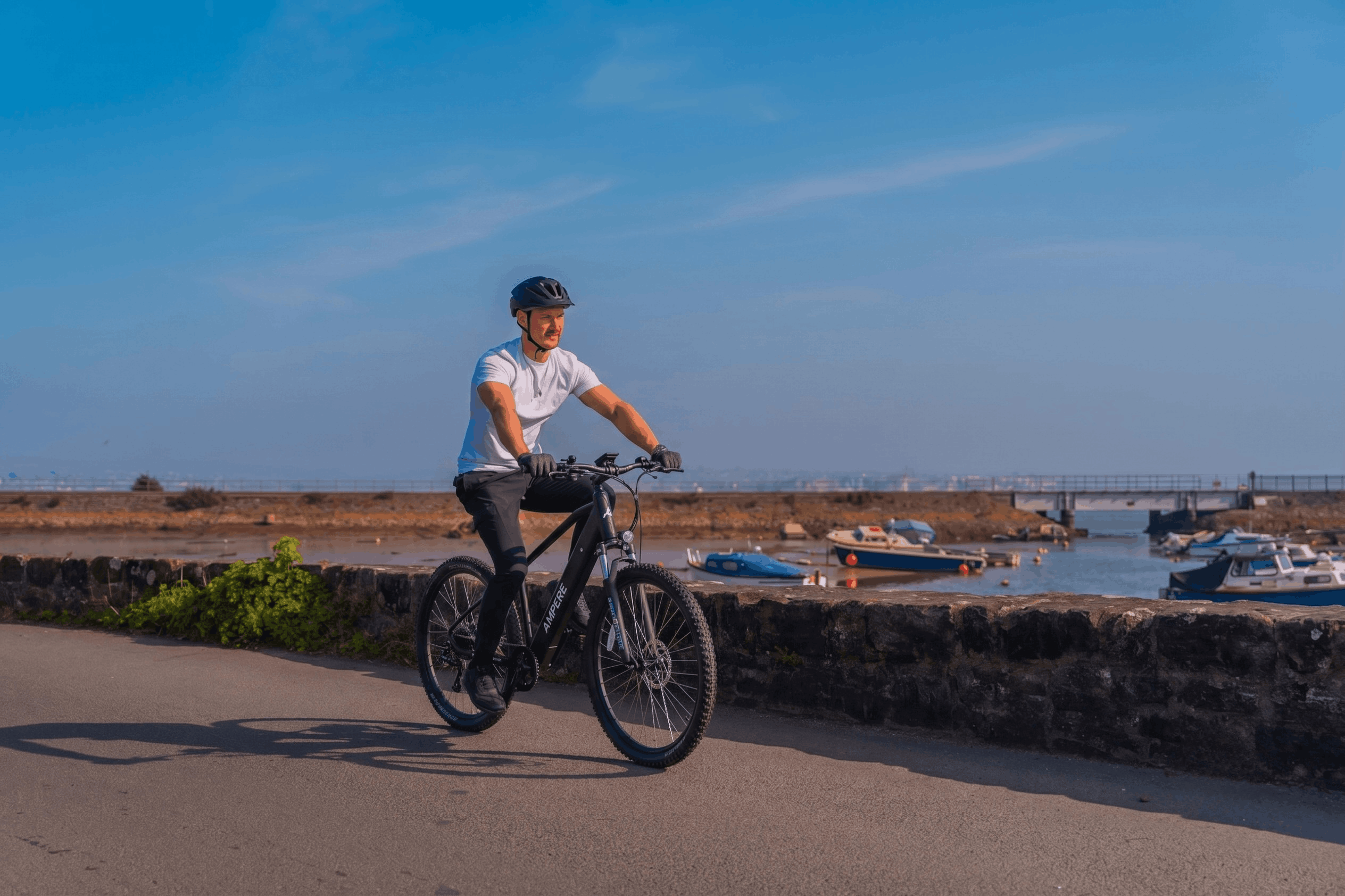 Black Ampere X-Trail electric mountain bike being ridden along a coastal path, highlighting its integrated battery, front suspension, hydraulic disc brakes, and rugged tires for mixed-terrain riding.